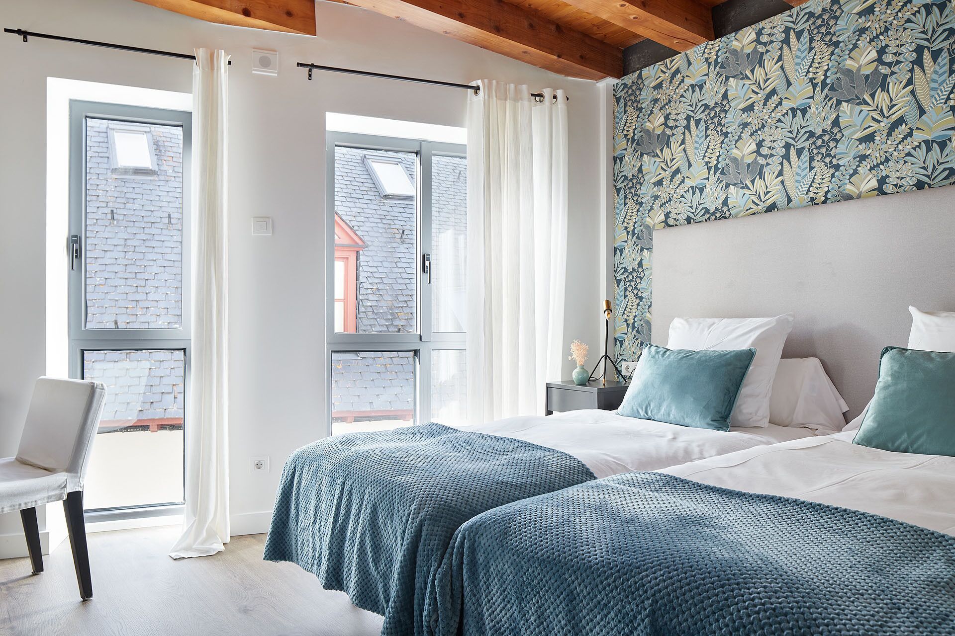 A bedroom with two beds, a chair, and a large window with curtains.