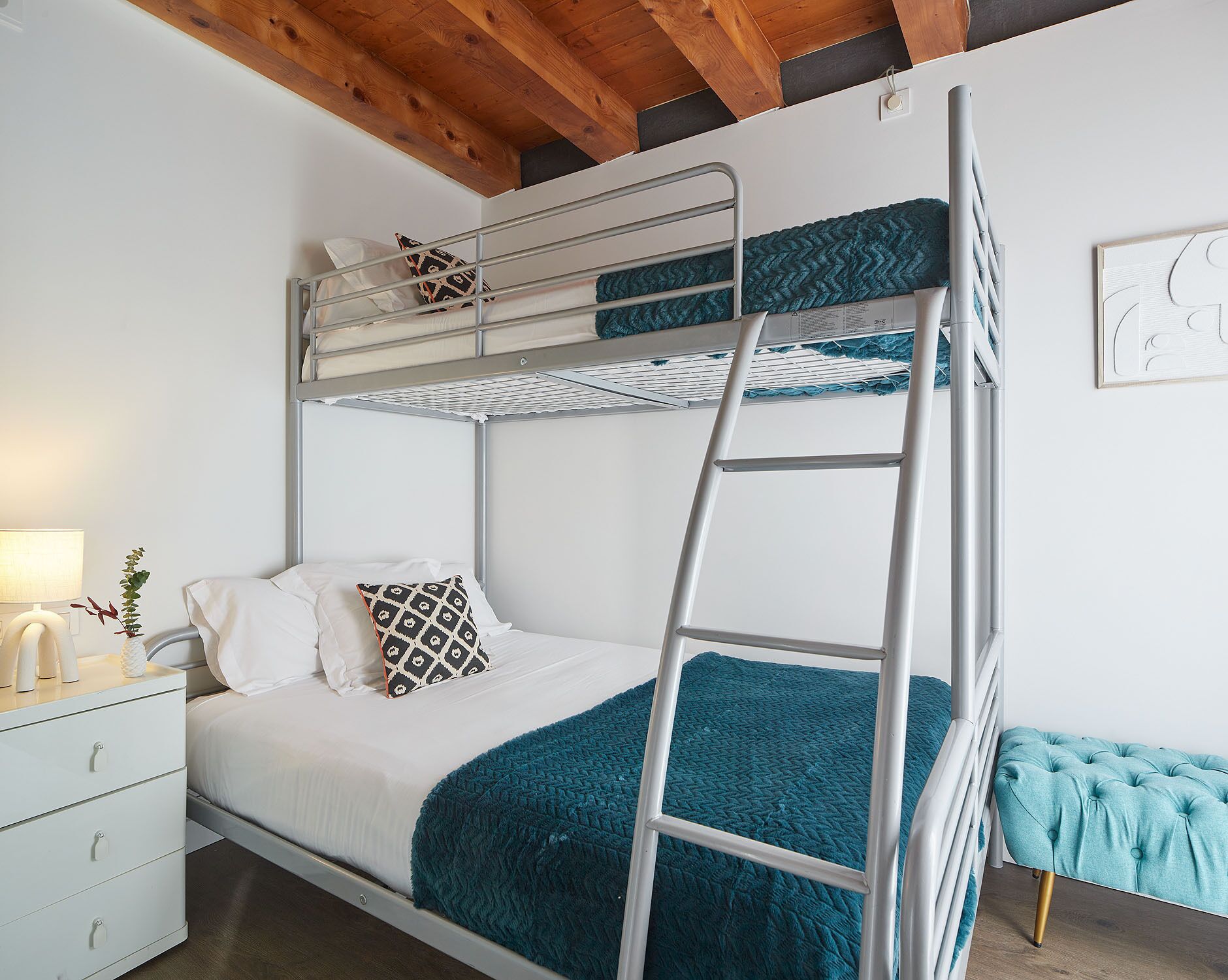 A bunk bed with a ladder, white bedding, and a teal blanket. There is a bedside table with a lamp and a plant.