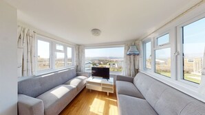 En rummelig stue med to grå sofaer, et fladskærms-tv på et hvidt skærmbord og store vinduer med udsigt over landskabet.