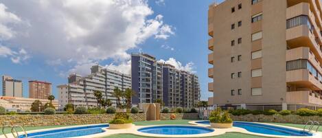 A modern apartment building with a pool area, including two swimming pools and a green lawn.