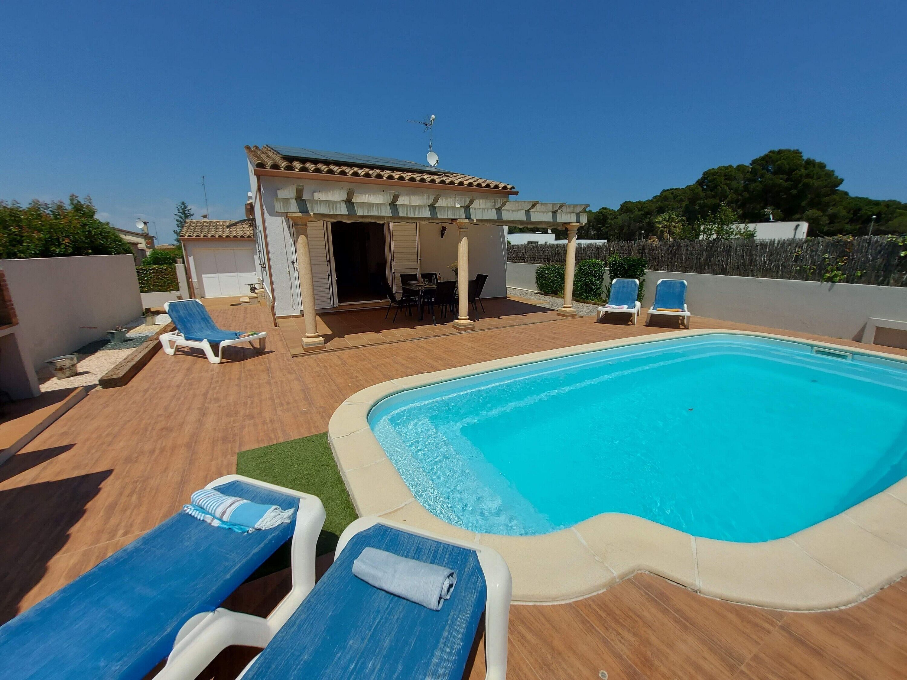 A swimming pool with lounge chairs, a covered patio area, and a small building with a tiled roof.
