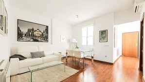 A modern living room with a white sofa, glass coffee table, and a cityscape painting. - Chueca Urban (Madrid)