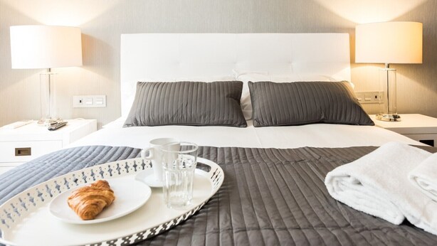 A neatly made bed with pillows, a tray with a croissant and glasses, and a folded towel on a bed with a grey quilt. - Chueca Urban (Madrid)