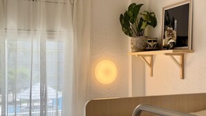 Une pièce avec une fenêtre, un luminaire fixé au mur, une étagère avec une plante et une maquette de petit véhicule, et une tête de lit en bois.