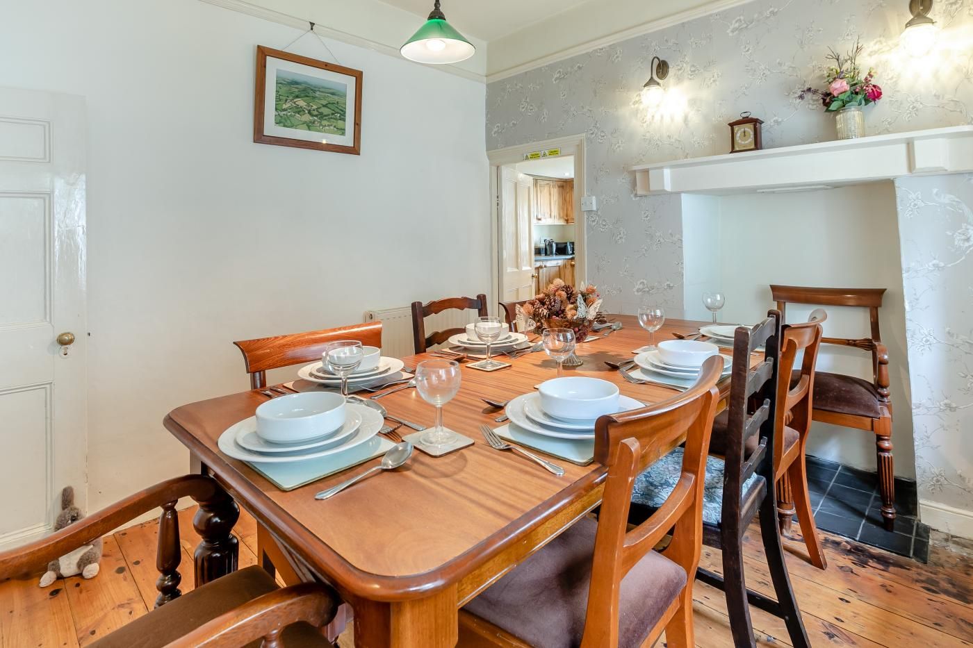 A dining room with a wooden table set for four, chairs, a framed picture on the wall, and a fireplace with a vase of flowers.