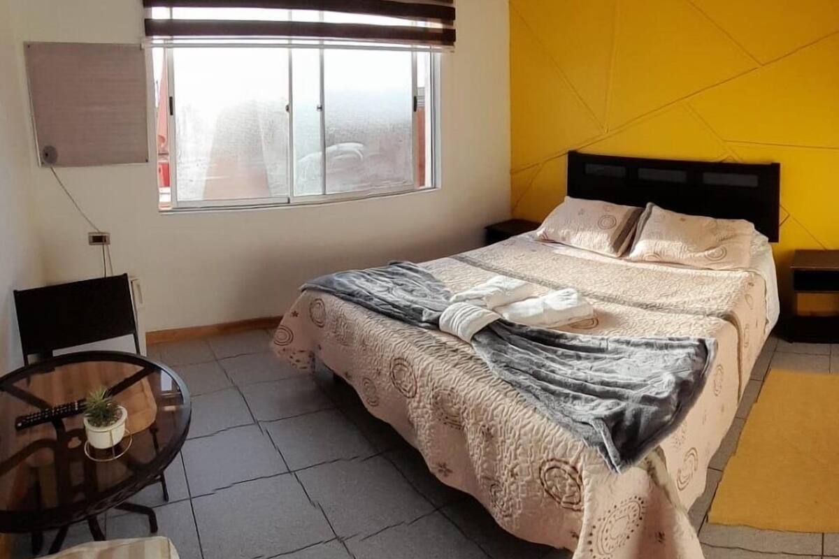 Ein Schlafzimmer mit Bett, einem Fenster mit Jalousien, einem Tisch mit Pflanze und einer gelben Akzentwand.