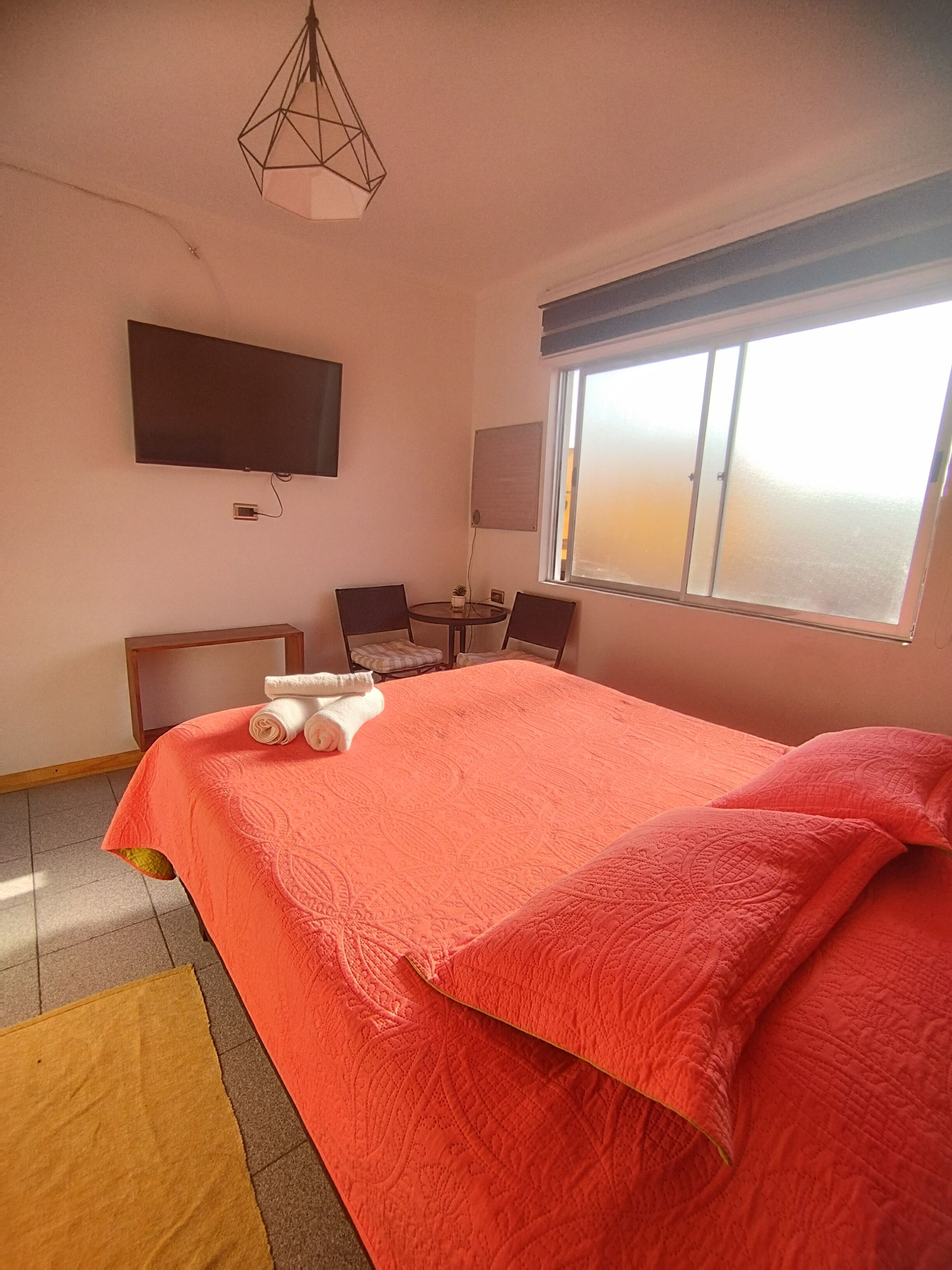 A hotel room with a large bed, a flat-screen TV mounted on the wall, a small table with chairs, and a window with blinds.