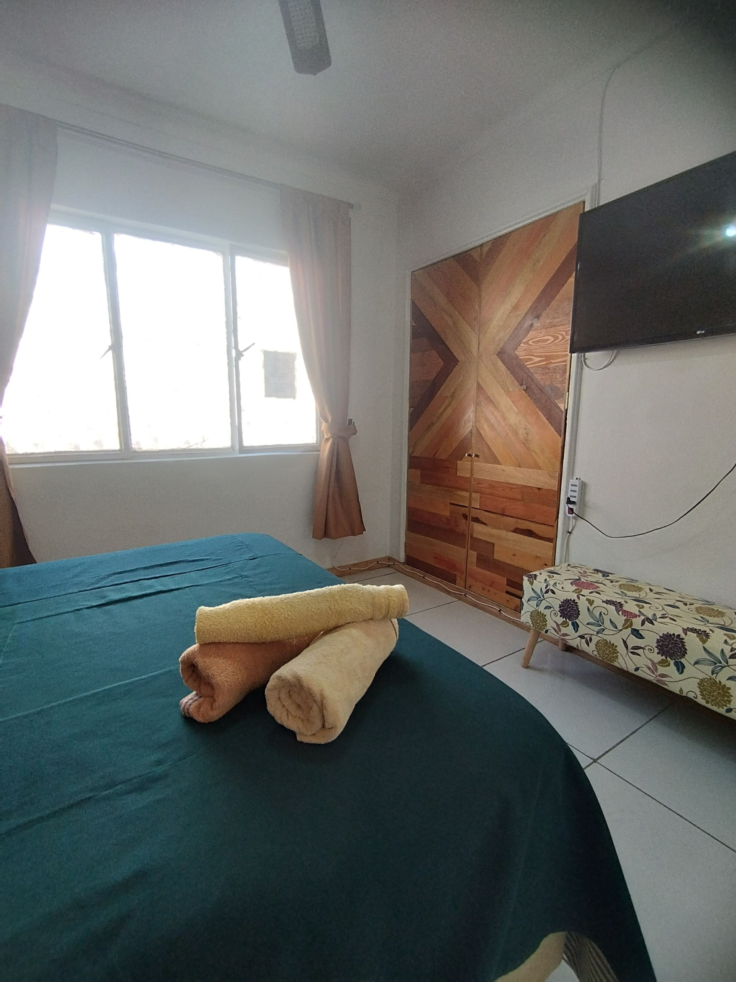 Una habitación con cama, televisor montado en la pared y ventana con cortinas.