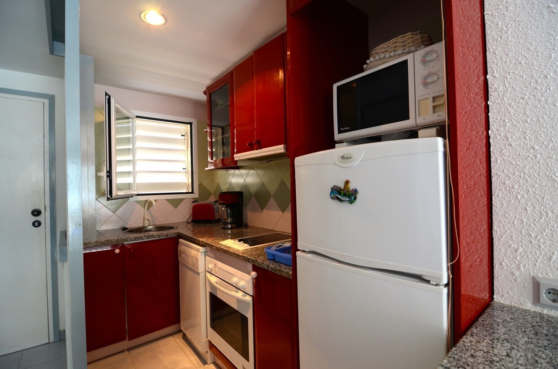A compact kitchen with red cabinets, a white refrigerator, a microwave, a sink, and a window with blinds.
