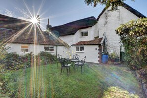 Uma casa branca com telhado de telhas, um jardim com mesa e cadeiras, e uma chaminé visível.