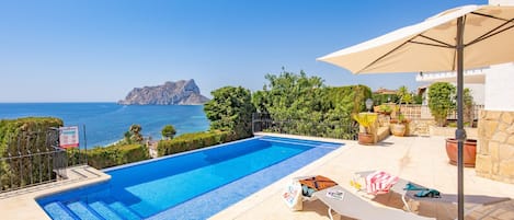 A swimming pool with a view of the sea and a rocky island, surrounded by greenery and outdoor seating.