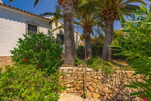 A white building with a stone base, surrounded by lush greenery and palm trees. - Villa Quimera - PlusHolidays (Benissa)