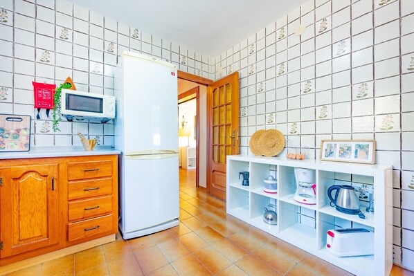 A kitchen with a refrigerator, microwave, wooden cabinets, and a white shelf unit with kitchen appliances. - Villa Quimera - PlusHolidays (Benissa)