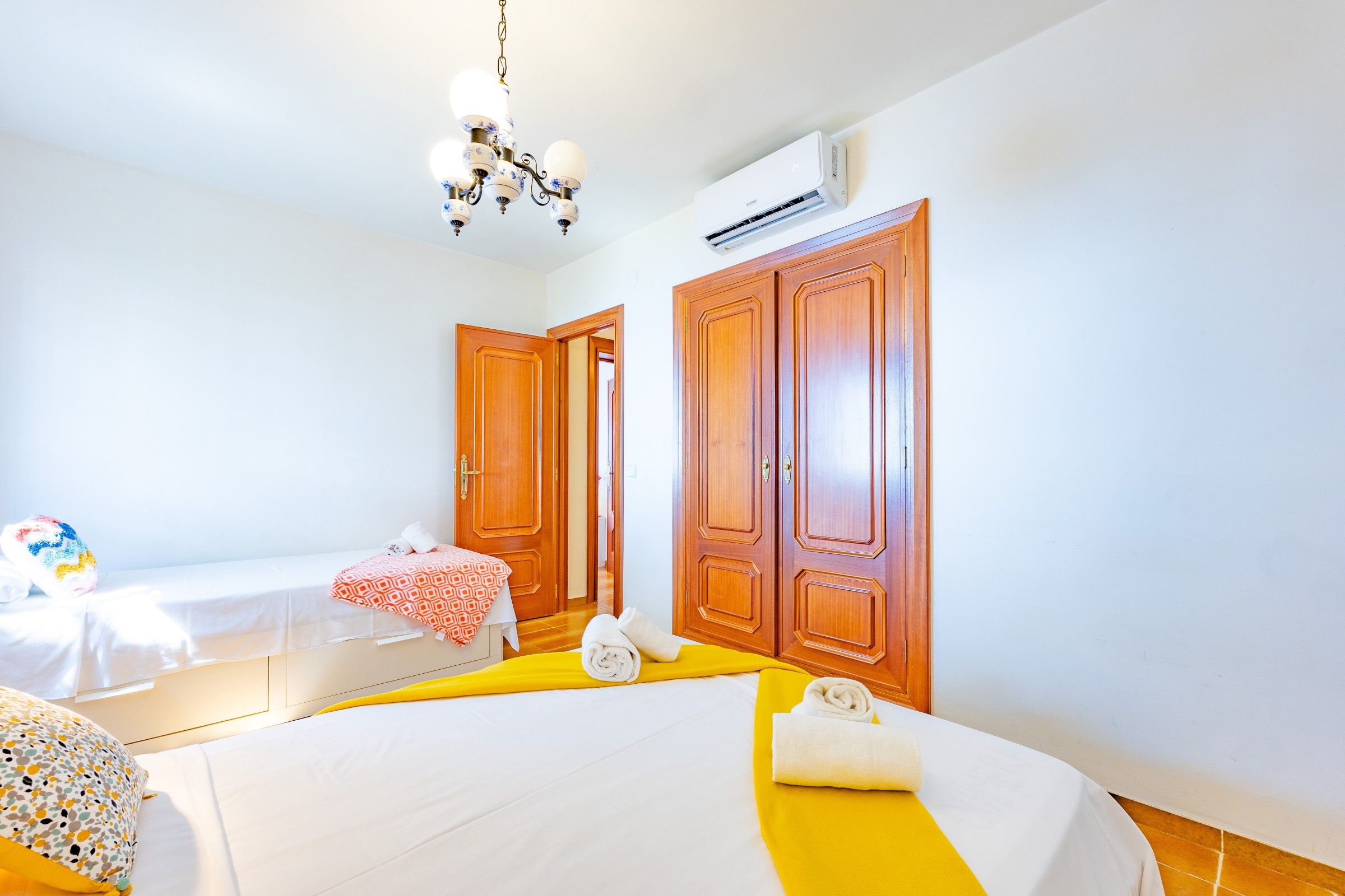 A hotel room with two beds, a chandelier, an air conditioning unit, wooden wardrobes, and a small table with a red and white tablecloth.