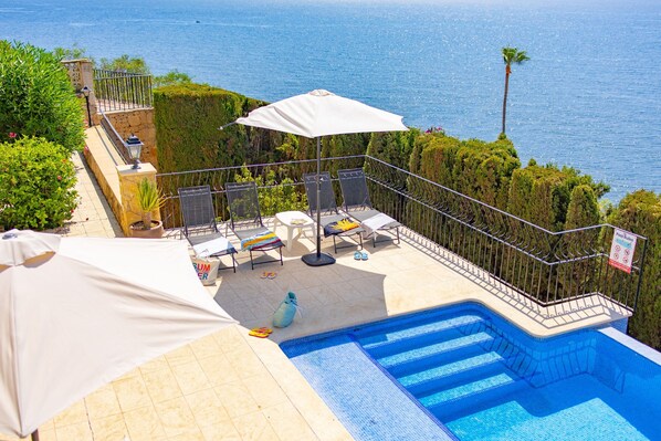 A poolside area with a view of the sea, a white umbrella, and lounge chairs. - Villa Quimera - PlusHolidays (Benissa)