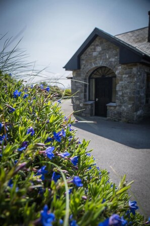 Un bâtiment en pierre avec une porte sombre, entouré de verdure et de fleurs bleues.