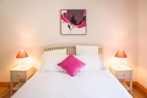 Un lit avec une literie blanche et un seul oreiller rose, flanqué de deux tables de chevet avec des lampes, dans une pièce dont le mur est uni et où un tableau encadré est accroché au-dessus du lit.