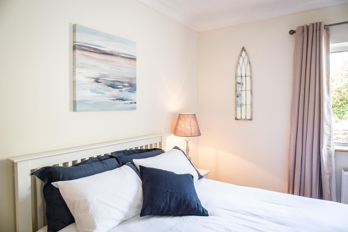 Un dormitorio con una cama, almohadas, una lámpara de pared, una ventana con cortinas y un cuadro.