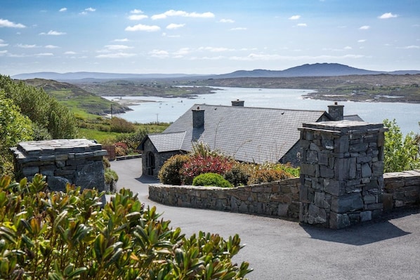 A stone building with a sloped roof, surrounded by a stone wall and lush greenery, overlooking a body of water and distant hills. - Once Upon a Tide (Clifden)