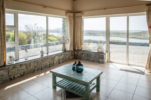 Une pièce avec un mur en pierre, une table en bois et de grandes fenêtres donnant sur un lac et des collines.