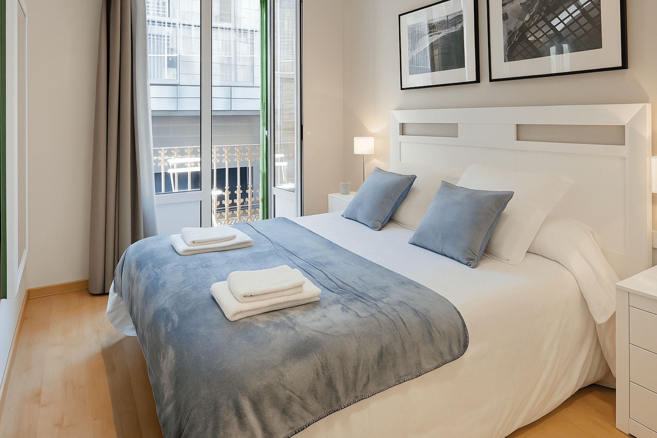 A neatly made bed with a grey and white duvet, two pillows, and folded towels. There is a window with a view of a building and a balcony.