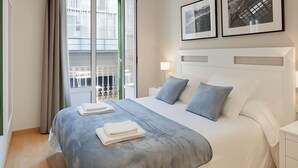 A neatly made bed with a grey and white duvet, two pillows, and folded towels. There is a window with a view of a building and a balcony.