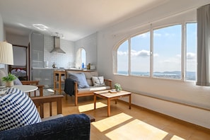 Un salon moderne avec un canapé, une table basse et une table à manger. On aperçoit une cuisine à l’arrière-plan, avec vue sur la ville.