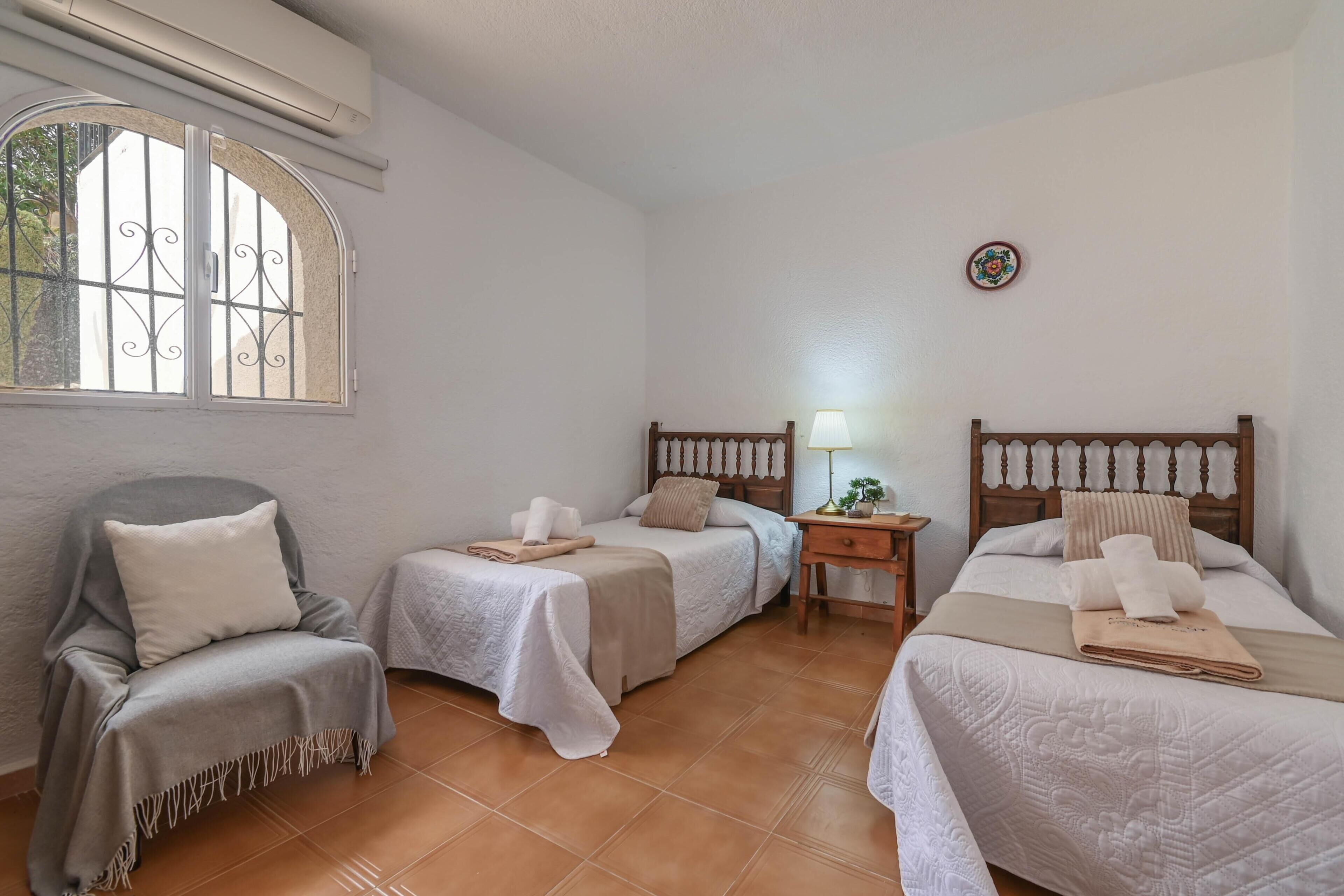 A room with three beds, a chair, a small table, a lamp, and a window with bars.