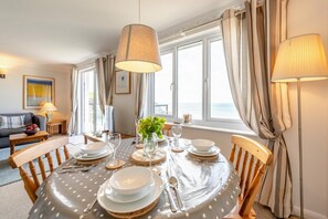 A dining area with a round table set for four, a vase with flowers, and a view of the sea through large windows. - 2 Bed in Putsborough (oc-flat31) (Croyde)