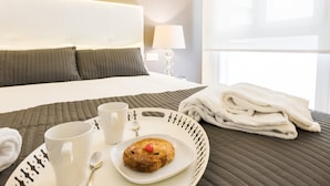 Ett hotellrum med en snyggt bäddad säng, ett bricka med ett bakverk och två muggar, och en hopvikt handduk på sängen.