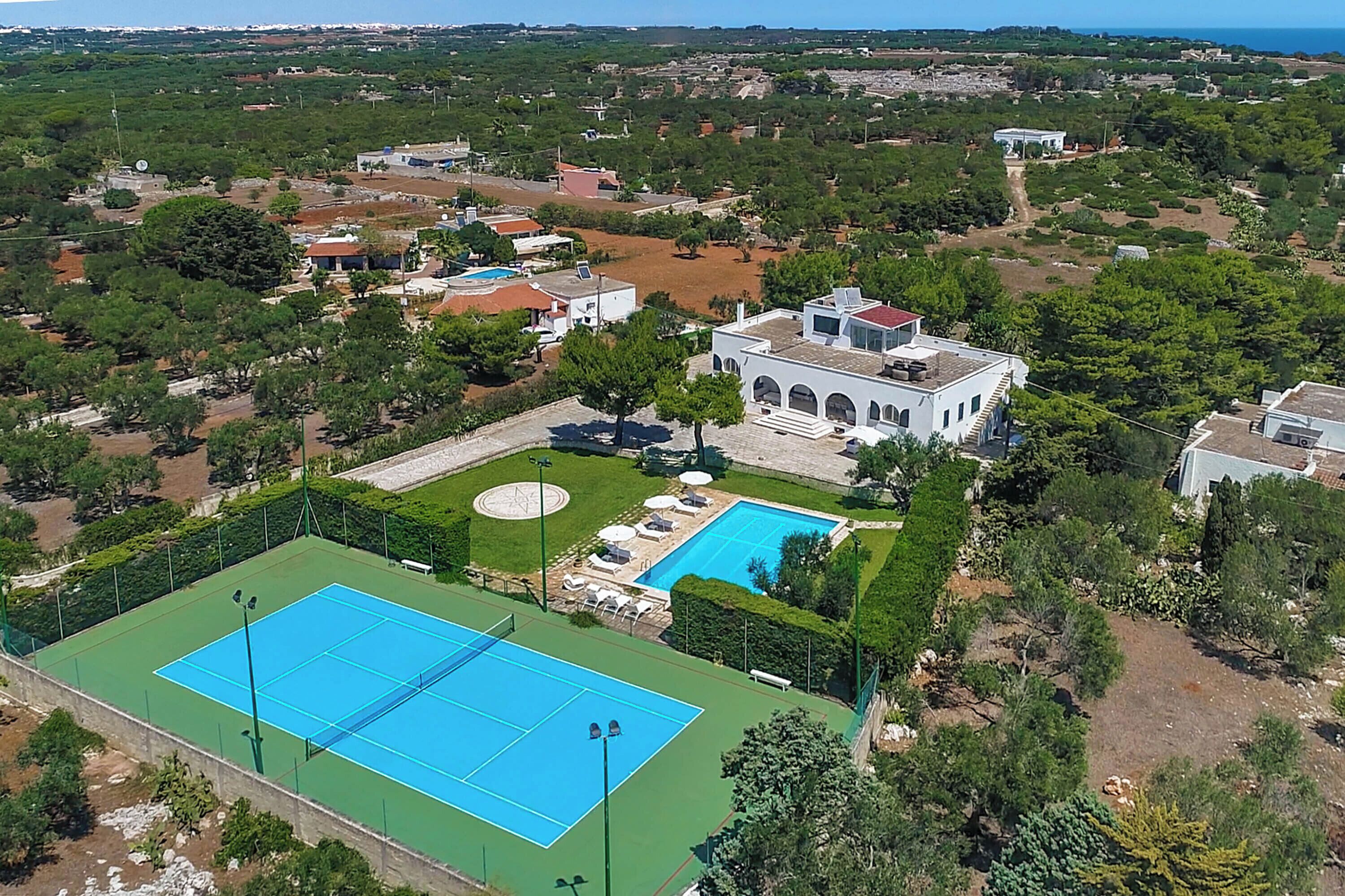 Vue aérienne d’un quartier résidentiel avec un court de tennis, une piscine et une grande villa dotée d’un jardin.