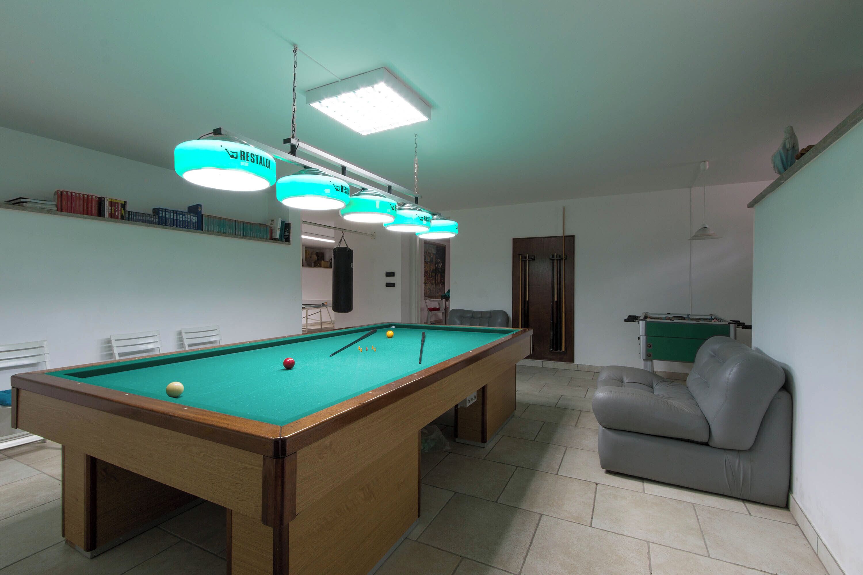 Una mesa de billar con superficie de fieltro verde, un sofá gris y una mesa de futbolín en una habitación con paredes blancas y suelo de baldosas.