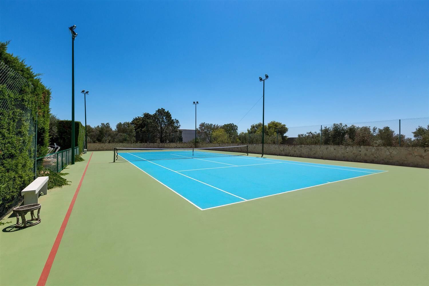 Cancha de tenis al aire libre con superficie verde, rodeada por una valla y árboles.