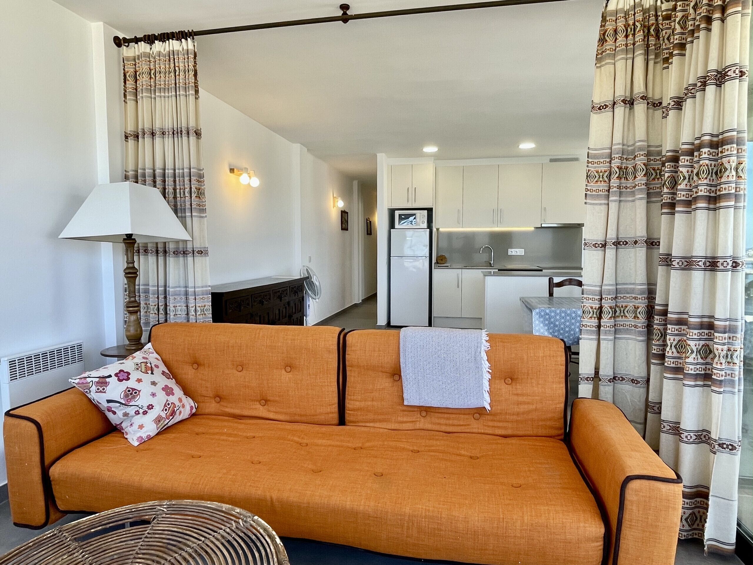 A living room with an orange sofa, a lamp, patterned curtains, a wicker chair, and a kitchen in the background.