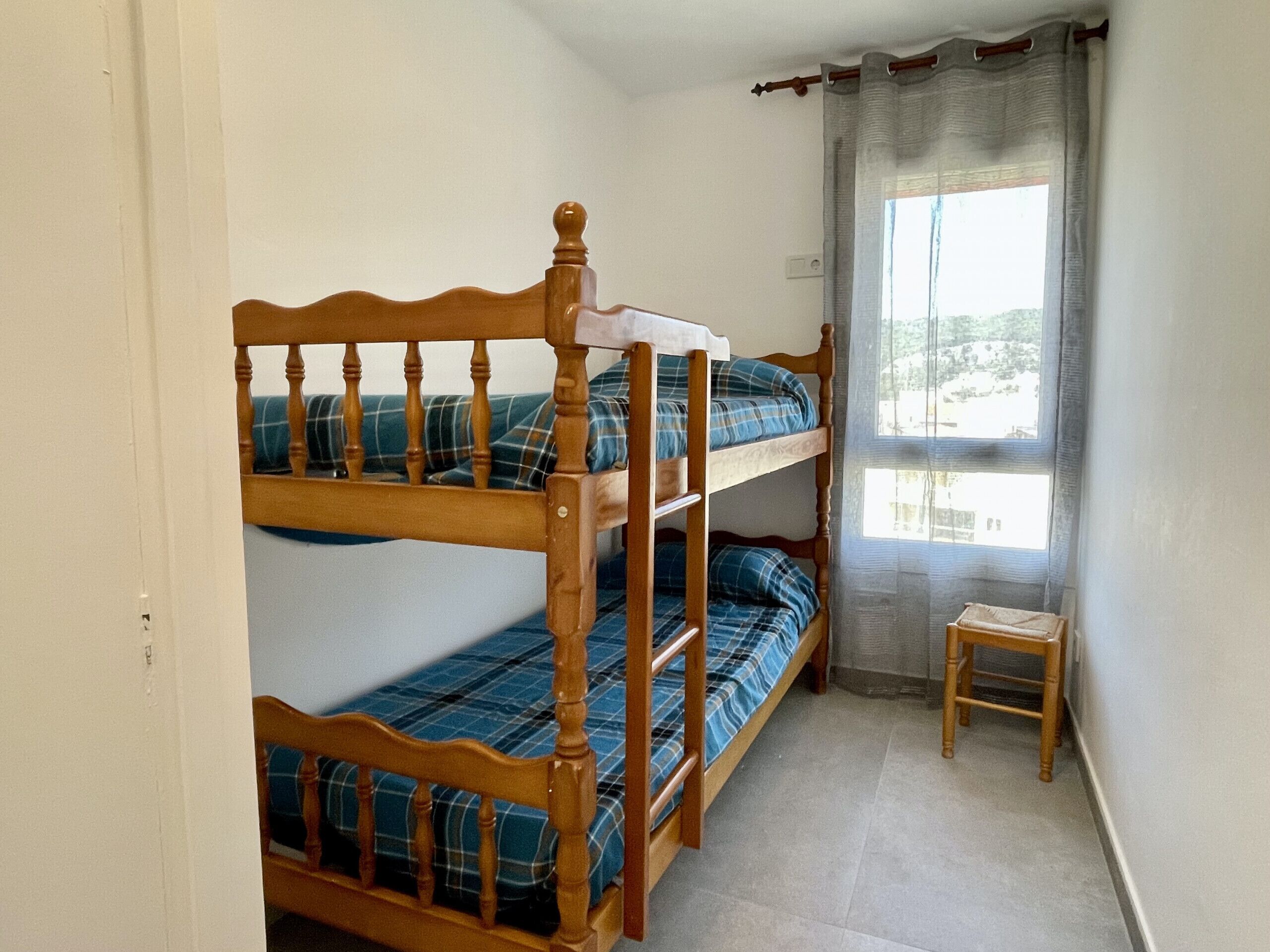 A bunk bed with a wooden headboard, a small wooden stool, and a window with curtains.