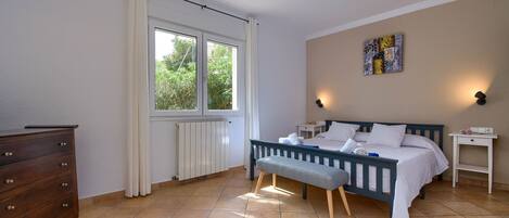 A bedroom with a bed, a dresser, a window with curtains, a radiator, and a wall-mounted light fixture.