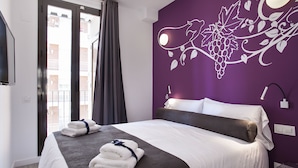 Hotellihuone, jossa on sänky, violetti seinä, jossa on viiniköynnöskuviota, ikkuna verhoilla ja näkymä ulkona oleviin rakennuksiin.