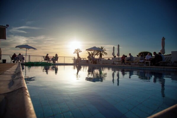 A poolside area with lounge chairs and umbrellas, a clear sky, and the sun setting in the background.
