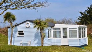 A small, light blue house with a white door and windows, a bench, and palm trees in the background. - F67 Ischia, Riviere Towans (Hayle)