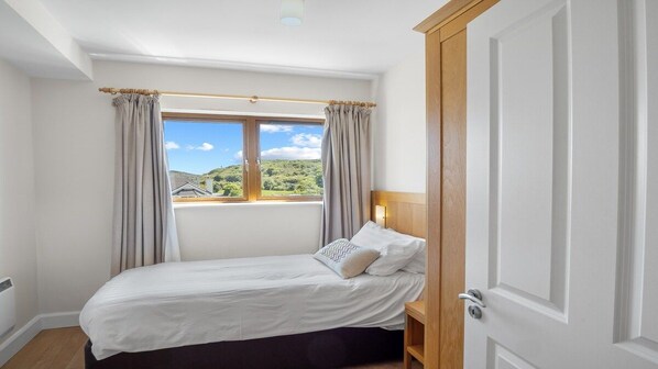 A bedroom with a bed, a window with curtains, a door, and a view of greenery outside.