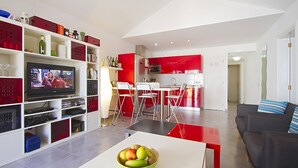 Un salon moderne doté d’un téléviseur à écran plat, d’un coin repas avec une table et des chaises rouges, et d’une cuisine avec des meubles de couleur rouge.