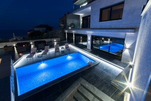 A modern outdoor swimming pool with lounge chairs and a glass-enclosed hot tub, surrounded by a contemporary house with a balcony.