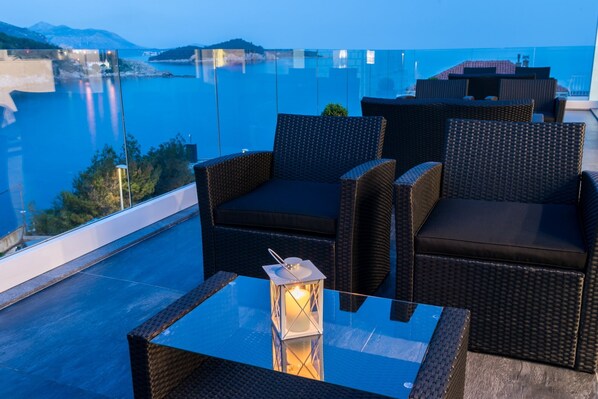 A balcony with wicker furniture and a glass table, overlooking a body of water and distant hills.