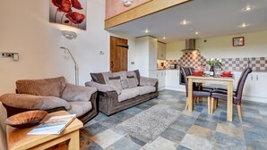 A cozy living room with a sofa and a dining table set for two, a kitchen in the background, and a checkered floor. - 2 Bed in Tiverton (oc-cide) (Cove)