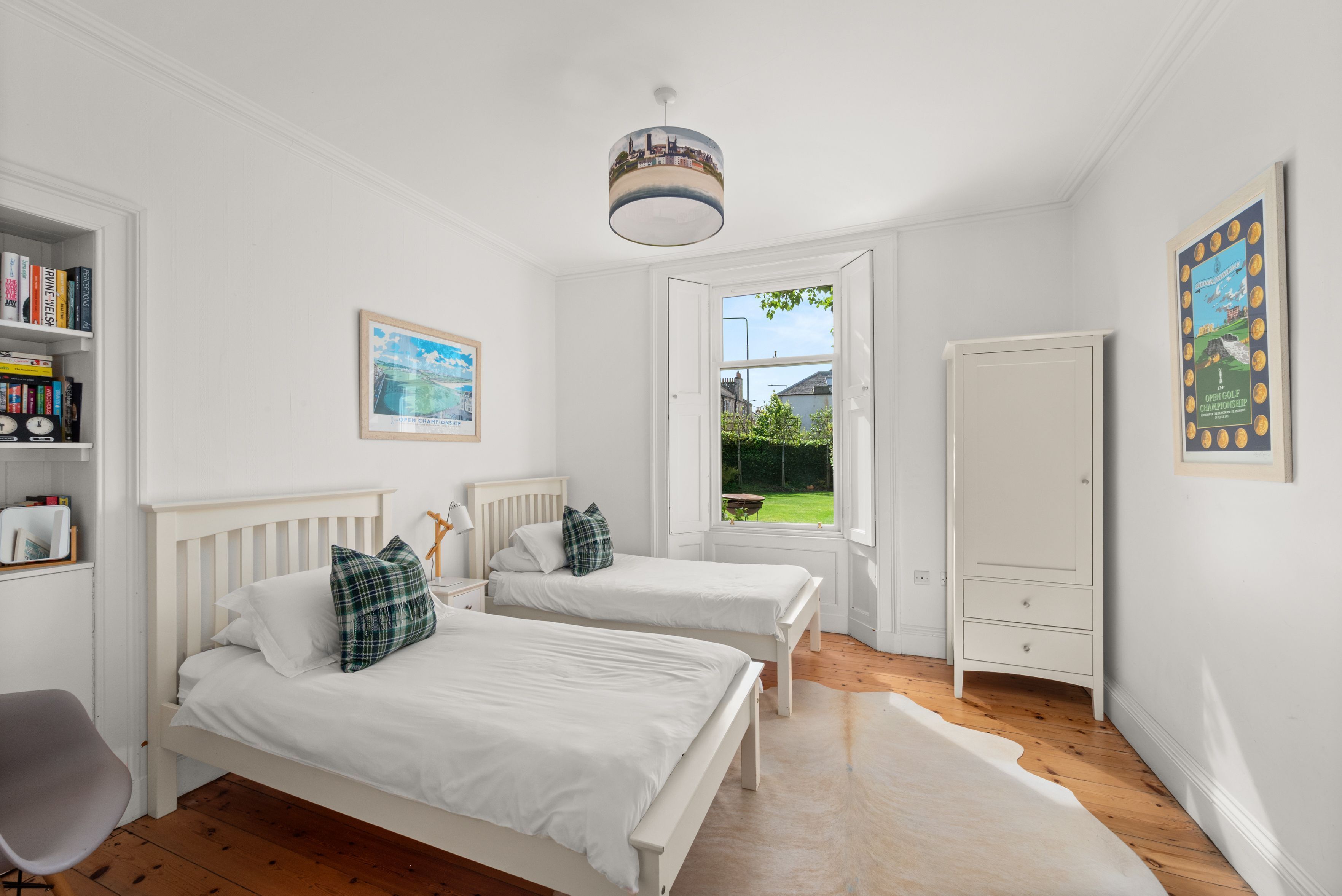 A bedroom with two single beds, a wardrobe, a bookshelf, and a window with a view of a garden.