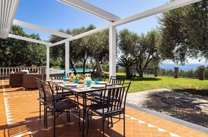 Un patio exterior con una mesa lista para comer, rodeado de sillas y vistas a una piscina y follaje.