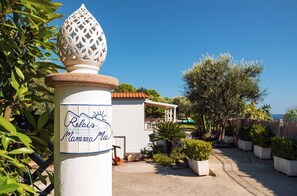 Opastekyltti, jossa lukee ”Relais Mamma Mia”, tiilikattoinen rakennus ja puutarha, jossa on ruukuissa kasvatettuja kasveja.