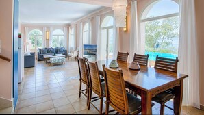 A spacious living area with a dining table, chairs, and a sofa. There are large windows with curtains, a TV, and a view of greenery outside. - Villa Agapi - Four Bedroom Villa, Sleeps 8 (Lefkada)