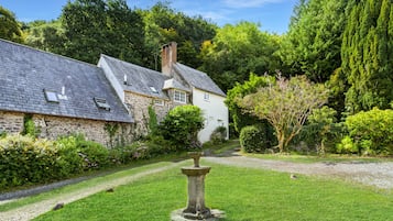 A stone building with a sloped roof, surrounded by lush greenery and a gravel path.