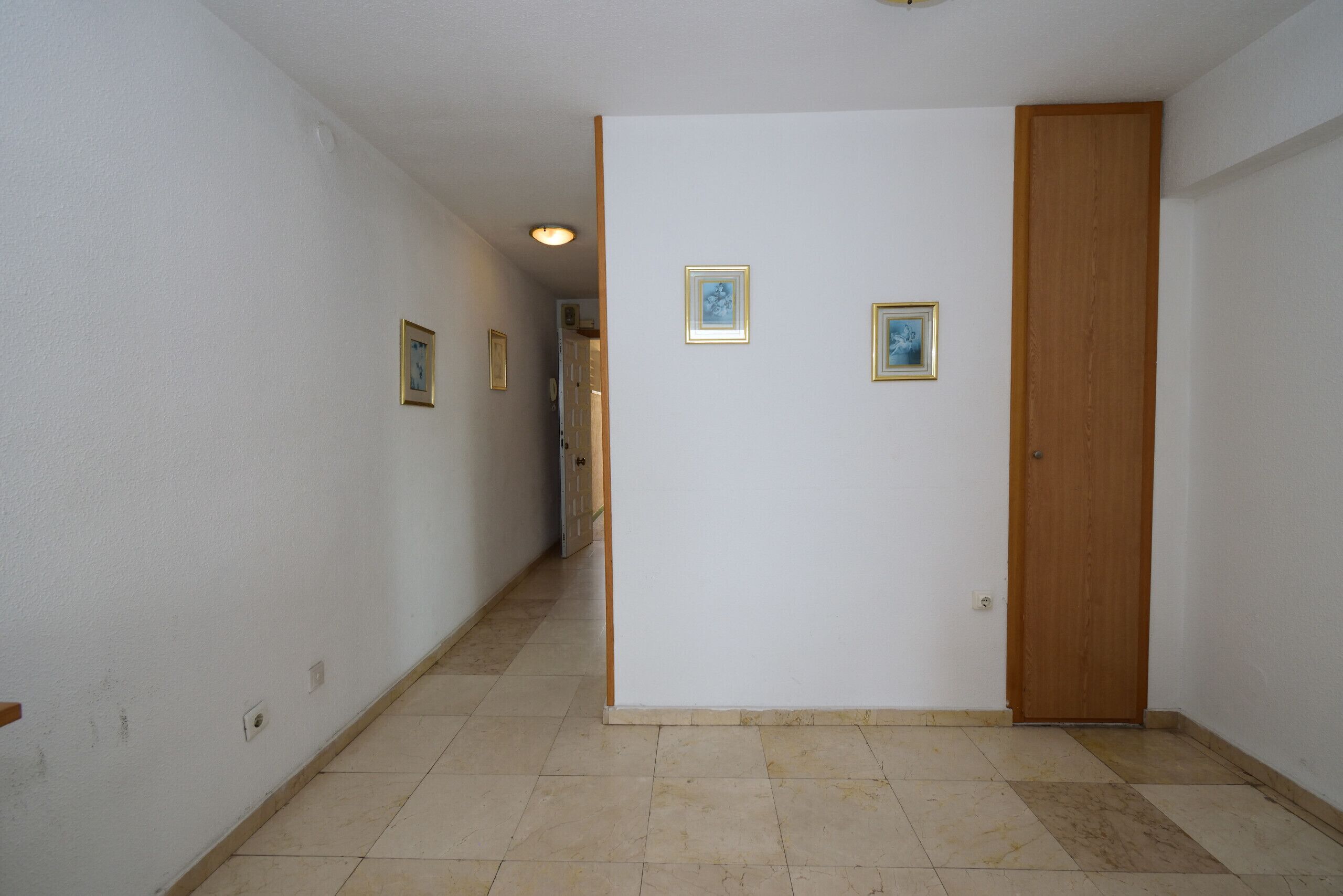 A hallway with white walls, a wooden door, and tiled flooring. There are two framed pictures on the wall.